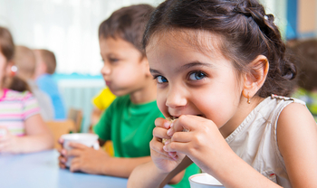 Ein kleines Mädchen mit langen braunen Haaren schaut fröhlich in die Kamera und beißt in ein Brot. Im Hintergrund sieht man noch weitere Kinder beim Essen und Trinken.