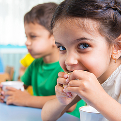 Ein kleines Mädchen mit langen braunen Haaren schaut fröhlich in die Kamera und beißt in ein Brot.