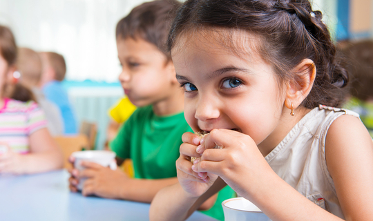 Ein kleines Mädchen mit langen braunen Haaren schaut fröhlich in die Kamera und beißt in ein Brot. Im Hintergrund sieht man noch weitere Kinder beim Essen und Trinken.