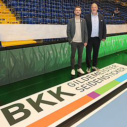 Zwei Personen stehen am Rand des Handballspielfeldes des TBV Lemgo Lippe, das BKK GS Logo ist im Fokus
