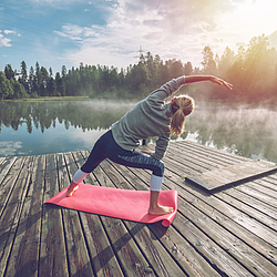 Junge Frau steht auf einer Yogamatte auf einem Steg am See und dehnt sich. Sie bewegt den linken Arm in die rechte Richtung. Das Wetter ist sonnig.