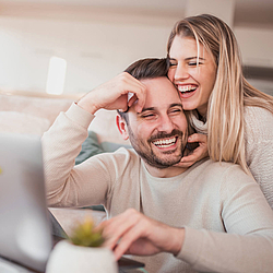 Ein Mann sitzt vor einen Laptop. Hinter ihn sitzt eine Frau, die ihn umarmt. Beide lachen.