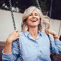 Eine ältere Frau mit langen grauen Haaren sitzt auf einer Schaukel. Sie trägt eine blaue Bluse und lacht ausgelassen.