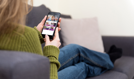 Eine Frau mit langen Haaren sitzt auf dem Sofa, man sieht über ihre Schulter auf ihr Handydisplay. Die Internetseite der BKK GS ist zu sehen.