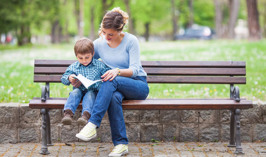 Eine junge Frau sitzt mit einem kleinen Jungen auf einer Bank. Sie lächelt und legt den Arm um den Jungen. Gemeinsam schauen sie sich ein Buch an.