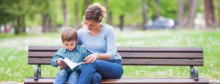 Mutter sitzt mit Kind im Arm auf einer Bank im Park und lesen zusammen ein Buch