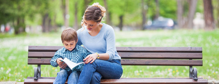 Stressbewältigung Mutter sitzt mit Kind im Arm auf einer Bank im Park und lesen zusammen ein Buch