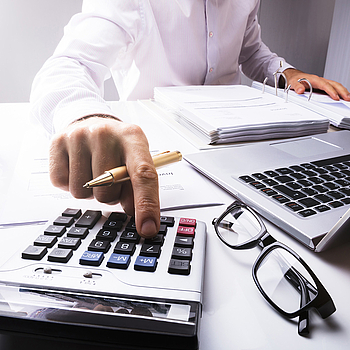 Ein Mann sitzt an einem Schreibtisch.Vor ihm liegen vielen Unterlagen und ein Laptop. Er tippt er etwas in einen Taschenrechner ein.