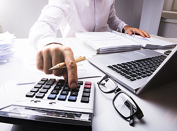 Ein Mann sitzt an einem Schreibtisch.Vor ihm liegen vielen Unterlagen und ein Laptop. Er tippt er etwas in einen Taschenrechner ein.