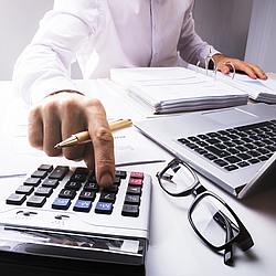 Ein Mann sitzt an einem Schreibtisch.Vor ihm liegen vielen Unterlagen und ein Laptop. Er tippt er etwas in einen Taschenrechner ein.