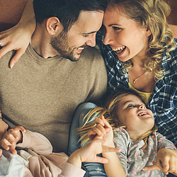 Eine junge vierköpfige Familie liegt zusammen auf dem Sofa. Die Frau umarmt ihren Mann und lacht ihn an.
