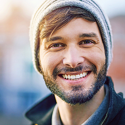 Ein Mann mit grauer Mütze und Bart lächelt.