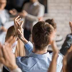 Eine Gruppe von Personen klatscht. Im Hintergrund sieht man einen Mann, der einer Frau etwas überreicht.