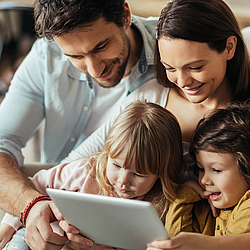 Eine junge Familie schaut gemeinsam auf ein Tablet. Die Mutter hält das Tablet fest, der Vater hat seine Arme um sie und die zwei Kinder gelegt.
