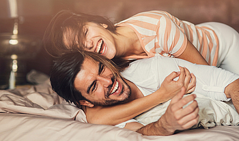 Eine junge Frau umarmt einen jungen Mann. Sie liegen im Bett und lachen ausgelassen.