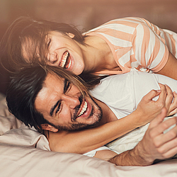 Eine junge Frau umarmt einen jungen Mann. Sie liegen im Bett und lachen ausgelassen.