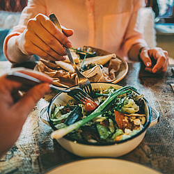 Zwei Personen sitzen an einem Tisch. Beide stechen mit der Gabel in die Schüssel mit Essen, die in der Mitte steht.