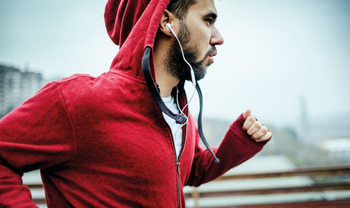 Ein Mann mit kurzen braunen Haaren und Bart trägt eine rote Sportjacke und weiße Kophörer. Er joggt.