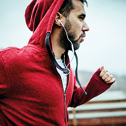 Ein Mann mit kurzen braunen Haaren und Bart trägt eine rote Sportjacke und weiße Kopfhörer. Er joggt.