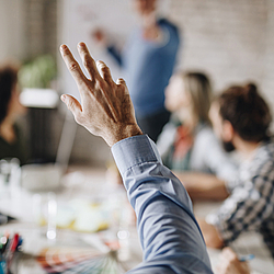 Eine Gruppe von Mitarbeitenden sitzt an einem großen Besprechungstisch. Ein Mann in einem blauen Hemd hebt seine Hand.. 