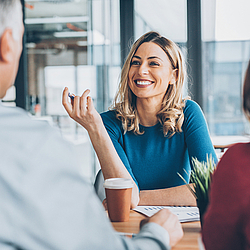 Eine blonde Frau sitzt einem Mann und einer Frau gegenüber. Sie unterhalten sich.