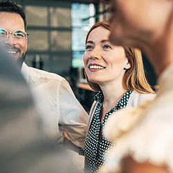 Eine Frau mit langen roten Haaren trägt eine dunkelgrüne Bluse mit weißen Pünktchen. Sie schaut lächelnd zu den Personen, die neben ihr stehen. 