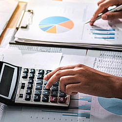 Es sind zwei Hände zu sehen. Die linke Hand bedient einen Taschenrechner. Die rechte Hand hält einen Kugelschreiber und zeigt auf ein Tortendiagramm auf einem Blatt Papier.