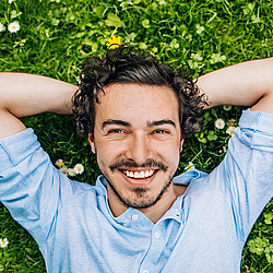 Ein junger Man mit lockigen schwarzen Haaren und Bart liegt auf einer Wiese voller Gänseblümchen. Er schaut lächelnd in die Kamera.
