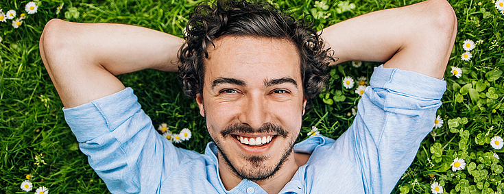 Ein junger Man mit lockigen schwarzen Haaren und Bart liegt auf einer Wiese voller Gänseblümchen. Er schaut lächelnd in die Kamera.