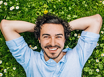 Ein junger Man mit lockigen schwarzen Haaren und Bart liegt auf einer Wiese voller Gänseblümchen. Er schaut lächelnd in die Kamera.