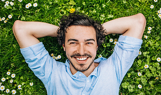 Ein junger Man mit lockigen schwarzen Haaren und Bart liegt auf einer Wiese voller Gänseblümchen. Er schaut lächelnd in die Kamera.