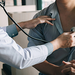 Eine Ärztin in einem weißen Kittel härt das Herz eines Patienten mit einem Stethoskop ab. Der Patient trägt ein blaues Hemd.