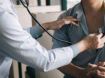 Eine Ärztin in einem weißen Kittel härt das Herz eines Patienten mit einem Stethoskop ab. Der Patient trägt ein blaues Hemd und hat die Finger ineinander verschränkt.