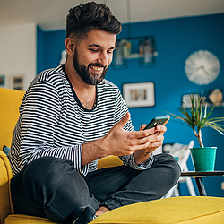 Ein Mann mit dunklen Haaren und Vollbart sitzt auf einem gelben Sessel und schaut lachend auf sein Smartphone.