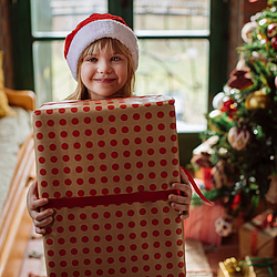 Ein Kind trägt eine Nikolausmütze und hält ein großes Geschenk in der Hand. Das Kind lächelt. Im Hintergrund steht ein Weihnachtsbaum.
