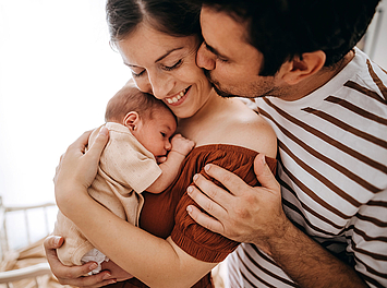 Eine Frau hält ihr neugeborenes Baby im Arm und lächelt. Ihr Partner steht hinter ihr und eine Hand auf ihre Schulter gelegt.