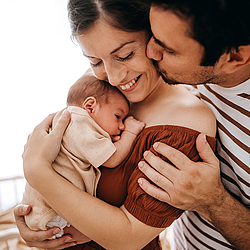 Eine Frau hält ihr neugeborenes Baby im Arm und lächelt. Ihr Partner steht hinter ihr und eine Hand auf ihre Schulter gelegt.
