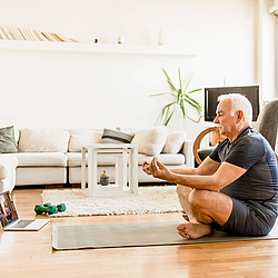 Ein Senior sitzt im Schneidersitz im Wohnzimmer auf einem Teppich. Er schaut dabei auf einen Laptop und führt eine Sportübung aus. 