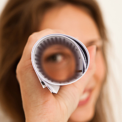 Eine Frau mit langen Haaren hält sich eine Rolle aus Papieren vor ein Auge und schaut lächelnd hindurch.