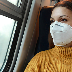 Frau mit FFP2-Maske sitzt im Zug und schaut aus dem Fenster.