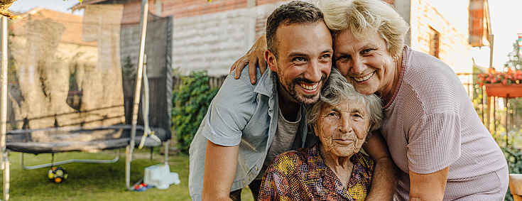 Eine Demenzpatientin sitzt im Garten auf einem Stuhl. Ihre Angehörigen, ein Mann und eine Frau, stehen hinter ihr und umarmen sie lachend.