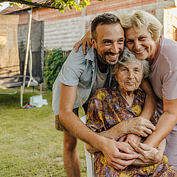Eine ältere Frau sitzt im Garten auf einem Stuhl. Ihre Angehörigen, ein Mann und eine Frau, stehen hinter ihr und umarmen sie.