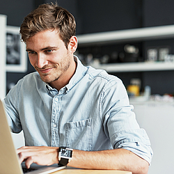 Ein Mann mit kurzen braunen Haaren sitzt in einer Küche am Tisch. Er tippt etwas auf der Tastatur seines Laptops und lächelt. 