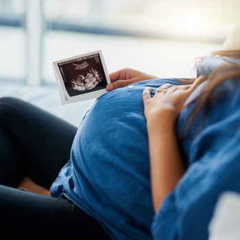 Eine junge schwangere Frau liegt auf dem Sofa. Ihre linke Hand liegt auf ihrem Bauch, in der rechten Hand hält sie ein Ultraschallbild ihres Babys.