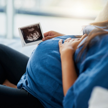Eine junge schwangere Frau liegt auf dem Sofa. Ihre linke Hand liegt auf ihrem Bauch, in der rechten Hand hält sie ein Ultraschallbild ihres Babys.