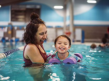 Eine Mutter ist mit ihrer kleinen Tochter in einem Schwimmbecken. Sie macht mit ihrer Tochter Schwimmübungen. Beide lächeln dabei.