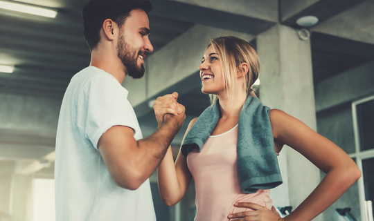 Ein junger Mann und eine junge Frau sind in einem Fitnessstudio und tragen Sportkleidung. Sie schauen sich an, lächeln und schlagen mit ihren rechten Händen ein.