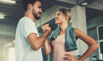 Ein junger Mann und eine junge Frau sind in einem Fitnessstudio und tragen Sportkleidung. Sie schauen sich an, lächeln und schlagen mit ihren rechten Händen ein.
