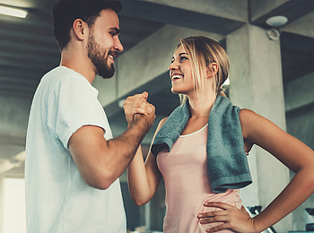 Ein Mann und eine Frau sind in einem Fitnessstudio und tragen Sportkleidung. Sie lächeln sich an und schlagen mit ihren Händen ein.