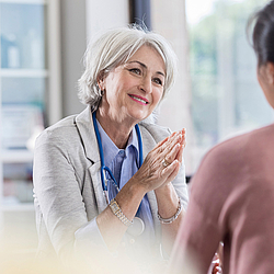 Eine ältere Hausärztin mit kurzen grauen Haaren führt ein Arztgespräch mit einer Patientin. Sie lächelt dabei freundlich.